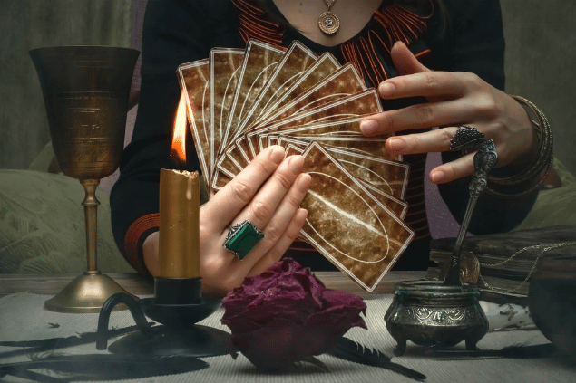 Tarot Cards Reading / Kreative Hub / Canva As mãos de uma taróloga segurando cartas de Tarot estão em destaque na imagem. Ao lado, há uma vela e outros objetos.
