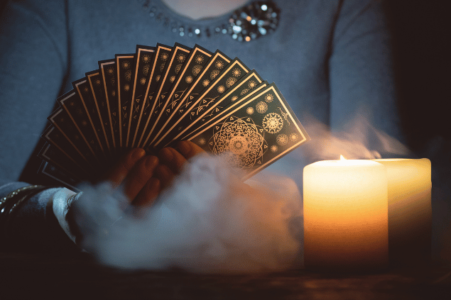 HappyNati / Getty Images / Canva As mãos de uma taróloga segurando cartas de Tarot estão em destaque na imagem. Há duas velas ao lado.