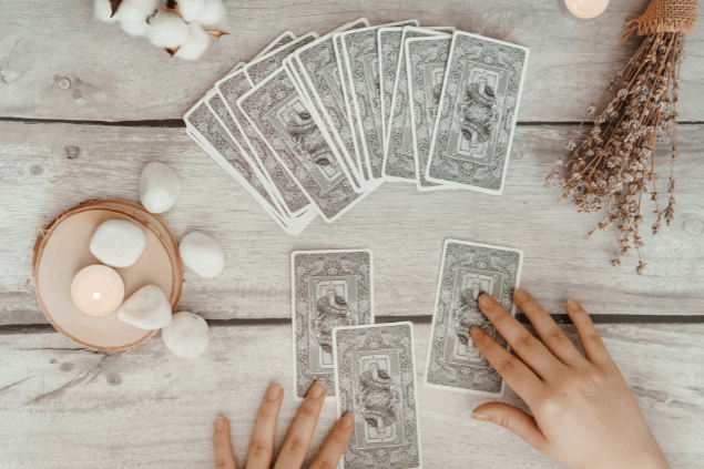 As mãos de uma taróloga posicionando cartas de Tarot viradas para baixo estão em destaque na imagem. Ao lado, há uma vela e outros objetos.
