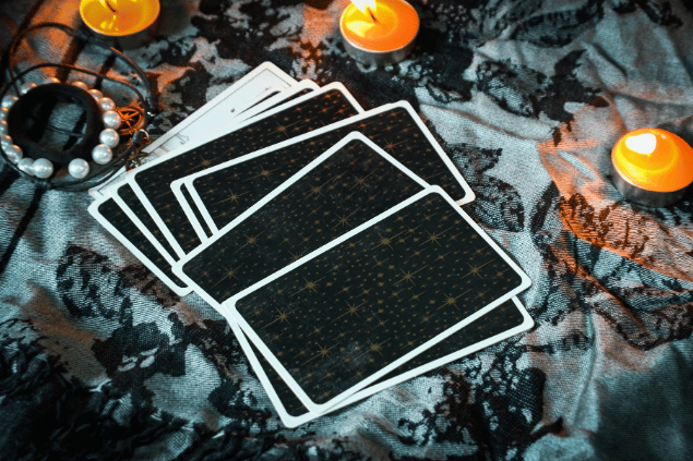 Bigc Studio / Canva Algumas cartas de Tarot viradas para baixo estão em destaque na imagem. Ao lado, há velas e outros objetos.