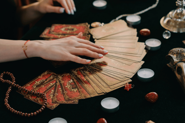 Mikhail Nilov / Pexels / Canva As mãos de uma taróloga posicionando cartas de Tarot viradas para baixo estão em destaque na imagem. Ao lado, há velas e pedras.