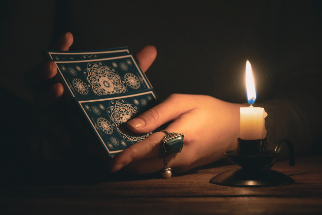 HappyNati / Getty Images / Canva As mãos de uma taróloga segurando cartas de Tarot viradas para baixo estão em destaque na imagem. Há uma vela ao lado.