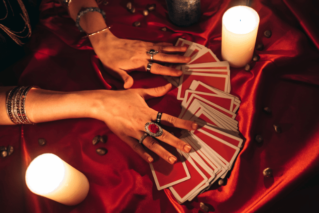 Urbazon / Getty Images Signature / Canva As mãos de uma taróloga sobre várias cartas de Tarot viradas para baixo estão em destaque na imagem. Há velas ao lado.