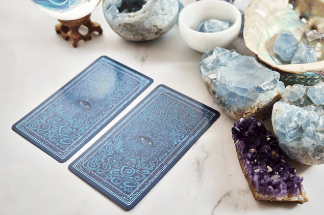 Vetrestudio / Canva Duas cartas de Tarot viradas para baixo estão em destaque na imagem. Ao lado, há alguns cristais e outros objetos.