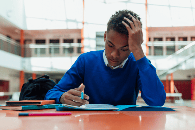 Menino negro concentrado enquanto estuda. Ele está sentado em uma mesa no pátio da escola.