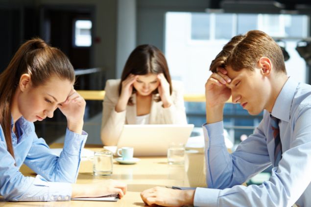 Imagem de três pessoas em um escritório com semblante de cansaço, com a cabeça baixa e com as mãos na testa