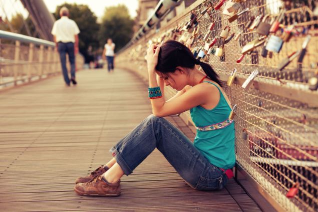 Imagem de uma mulher sentada no chão de uma ponte e está com a cabeça baixa e triste