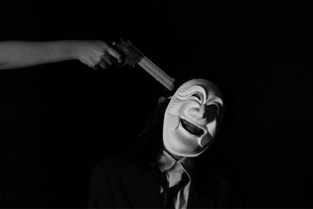 imagem de apenas o braço de uma pessoa com a arma na mão apontando para a cabeça de uma outra pessoa de máscara de palhaço