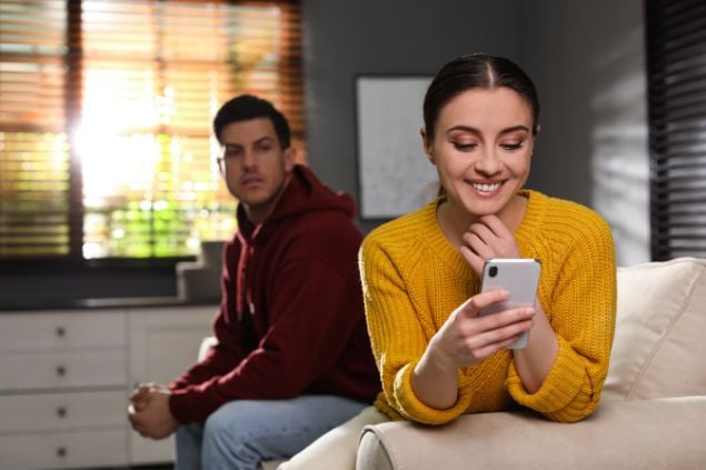 Homem olhando desconfiado para sua mulher que está sorrindo ao olhar para o celular
