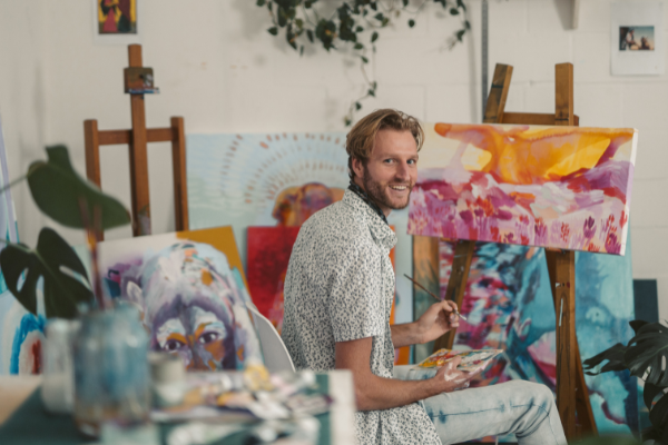 Homem olhando para o lado e sorrindo segurando pincel em uma sala cheia de quadros pintados