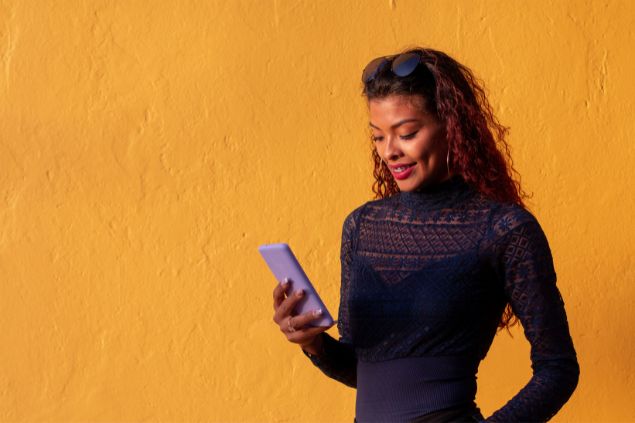 Imagem de uma mulher de roupa preta olhando o celular em um fundo amarelo