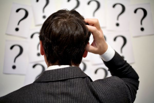 Homem de costas coçando a cabeça e olhando para diversos papéis com pontos de interrogação