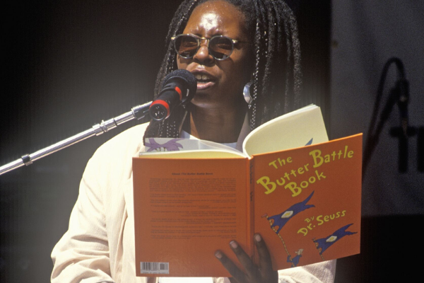 Whoopi Goldberg lendo um livro no palco com um microfone a sua frente