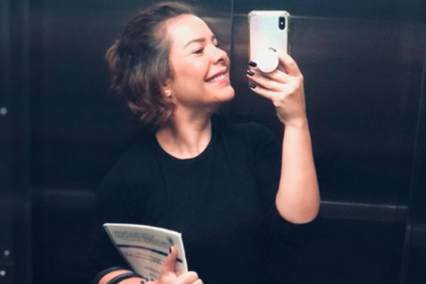 Fernanda Souza tirando selfie no espelho com seu celular enquanto segura sua bolsa e caderno