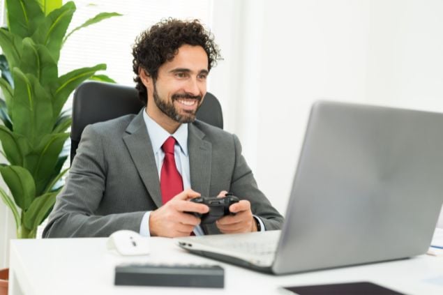 Imagem de um homem com um controel de video-game na frente de seu laptop, de terno, como se estivesse jogando na hora do trabalho