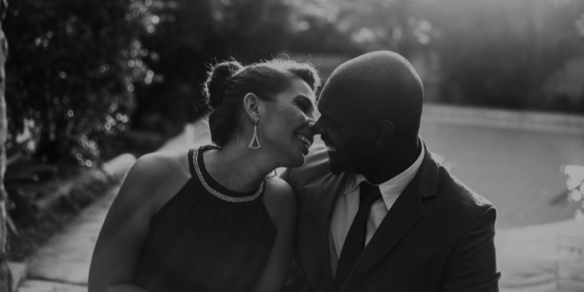 Um casal sorri e aproxima os rostos um do outro. A imagem é em preto e branco e ambos estão em um ambiente externo.