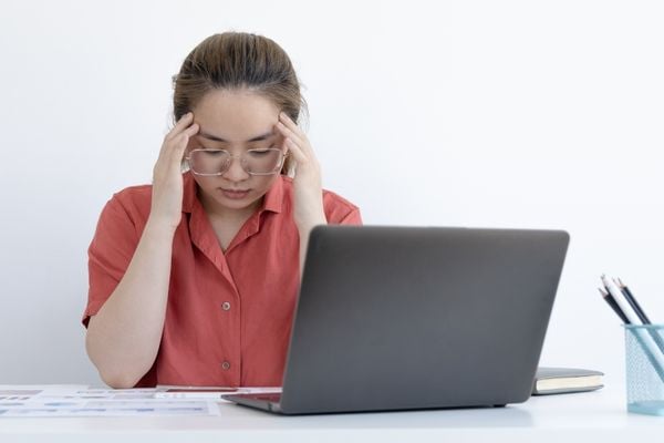 Mulher de óculos sentada na frente do notebook com as mãos na cabeça, em sinal de cansaço