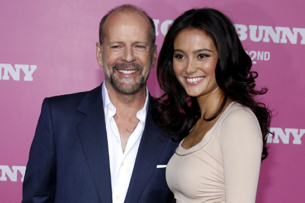 Bruce Willis sorrindo usando roupas sociais ao lado de uma mulher 