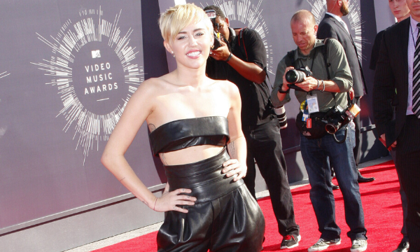 Miley Cyrus  no tapete vermelho  com as mãos na cintura e sorrindo com fotógrafos atrás