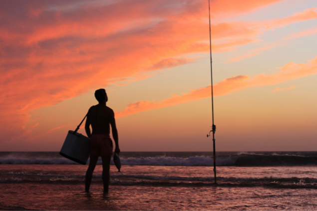 Silhueta de uma pessoa na beira da praia com uma bolsa no ombro, olhando o horizonte