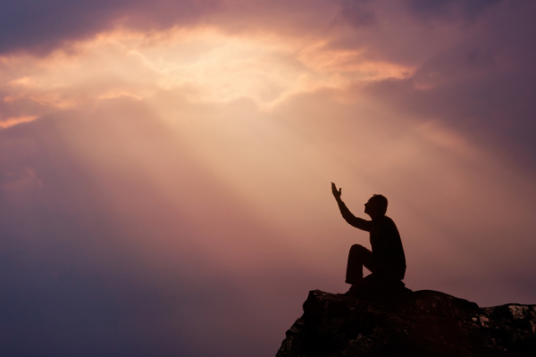 Homem sentado no topo de uma montanha com a mão levantada para o céu