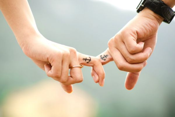 Dedos indicadores de duas pessoas entrelaçados. As duas mãos possuem uma tatuagem igual de âncora