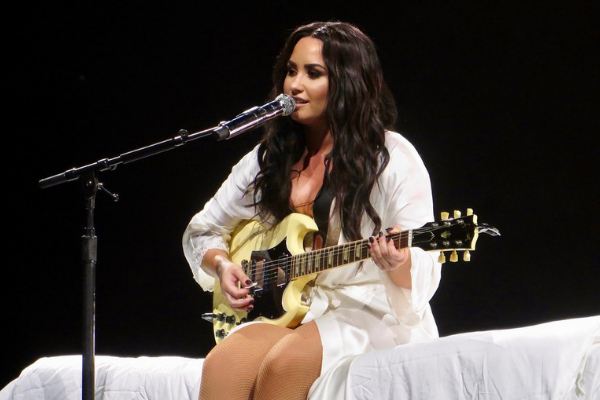 Imagem da cantora Demi Lovato sentada durante show, cantando e tocando uma guitarra
