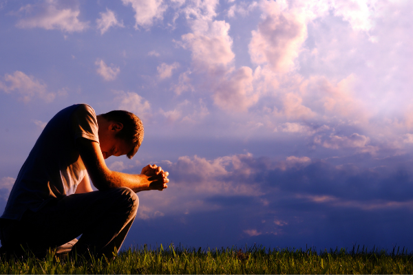 Homem sentado em um campo com as mãos fechadas e cabeça abaixada, em sinal de oração