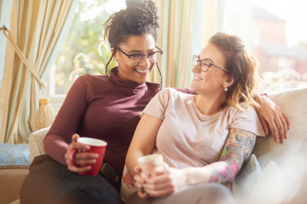 Duas mulheres sentadas no sofá com uma xícara na mão, sorrindo uma para a outra