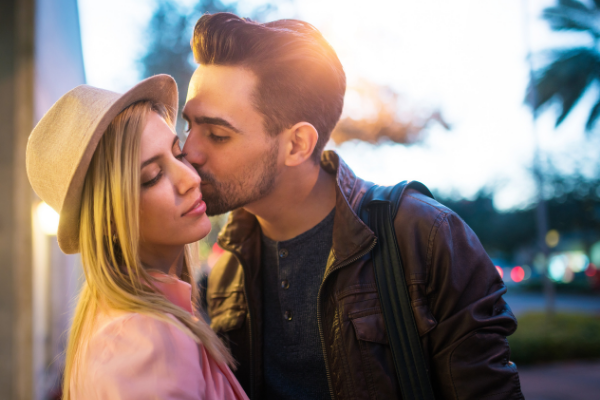Homem beijando uma mulher no rosto. A mulher usa um chapéu e ambos estão em um local público