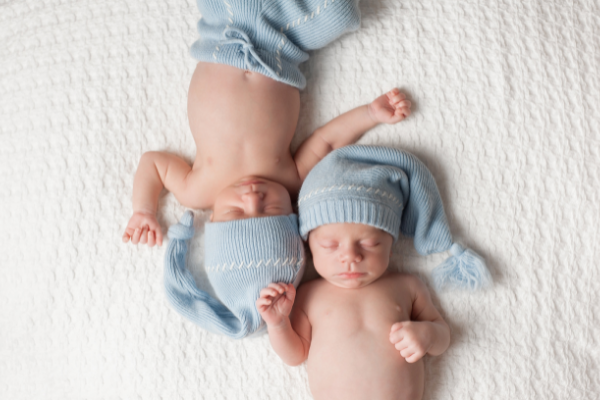 Imagem de gêmeos recém-nascidos deitados em uma cama e usando gorro