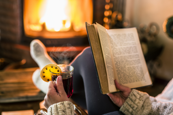 Pessoa lendo um livro na frente de uma lareira e tomando vinho quente