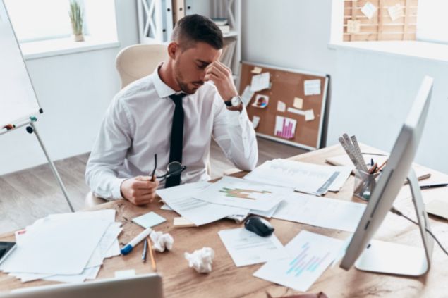 Homem exausto sentado em frente de uma mesa cheia de papéis e um computador