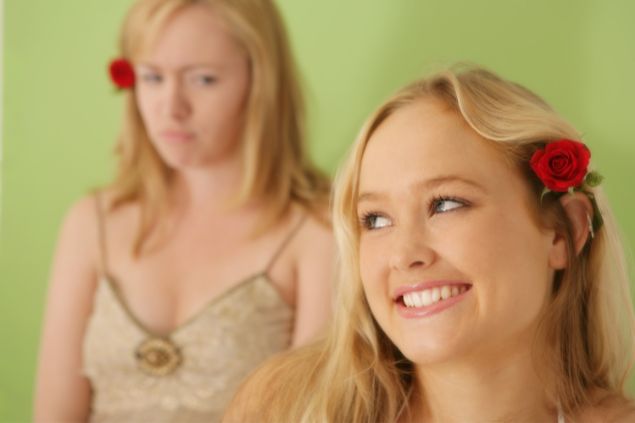 hidesy de Getty Images Signature/ Canva Imagem de uma moça sorrindo com uma flor atrás da orelha e outra olhando para ela com olhar de inveja