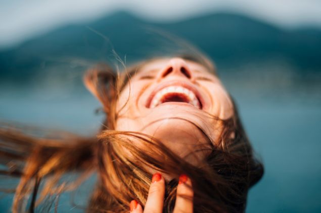 Imagem de uma mulher sorrindo com os olhos fechados e cabelos ao vento, ao ar livre