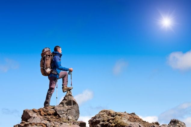 Imagem de um homem que escalou e chegou até o topo de uma montanha, onde é possível ver uma paisagem de céu azul