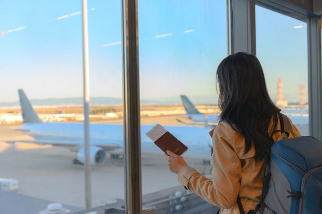 Imagem de uma mulher olhando pela janela em um aeroporto com a passagem de avião na mão e a mochila nas costas