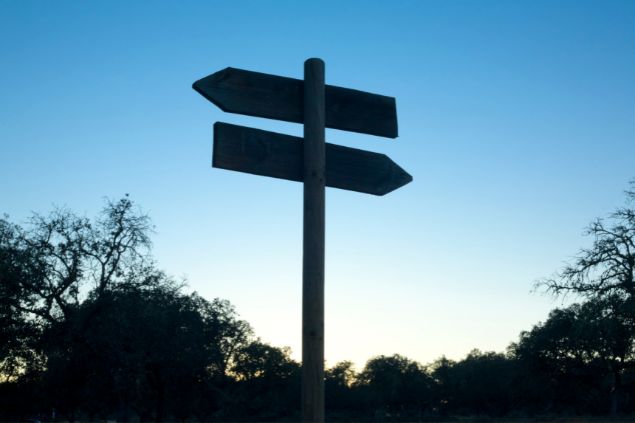 Imagem de uma placa indicando duas direções e atrás o céu azul