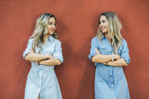 Imagem de duas irmãs gêmeas adultas, olhando uma para outra e sorrindo, encostadas em uma parede