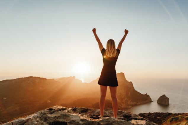 Imagem de uma mulher no topo de uma montanha, com os braços erguidos como se estivesse comemorando ao pôr do sol