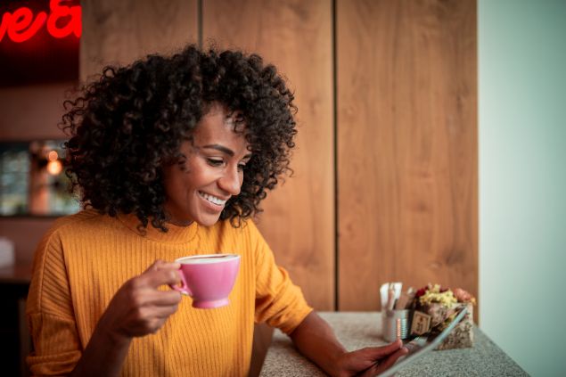 Imagem de uma mulher sorrindo enquanto toma café e olha o celular
