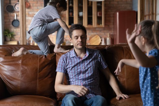 Imagem de um homem sentado no sofá, impaciente e seus filhos fazendo bagunça