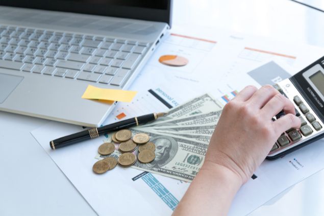 Imagem de uma pessoa fazendo cálculos com uma calculadora, em cima da mesa tem dólares, moedas e um notebook