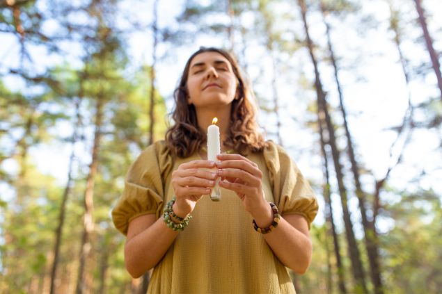 Imagem de uma mulher no meio da floresta com os olhos fechados e segurando uma vela