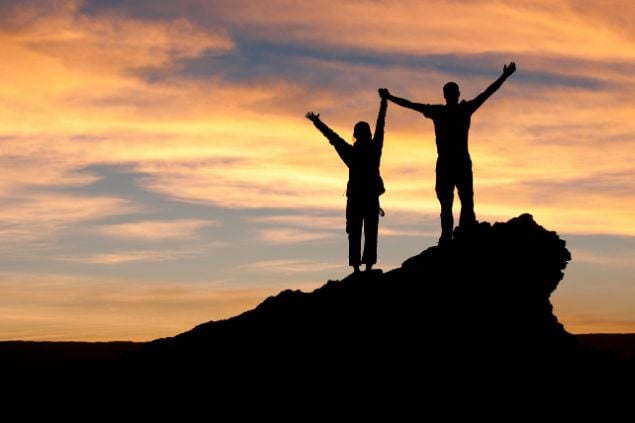 Imagem de duas pessoas com as mãos dadas e erguidas em sinal de comemoração no topo de uma montanha ao pôr-do-sol