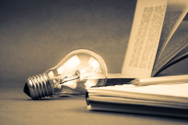 Imagem de um livro, um caderno e uma caneta em cima dele e uma lâmpada do lado