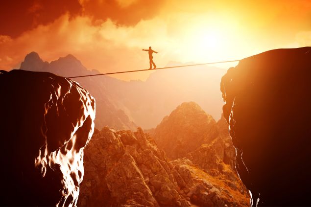 Imagem de uma pessoa atravessando em uma corda de uma montanha a outra e o pôr do sol atrás dela