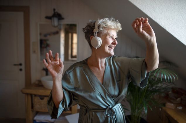 Imagem de uma senhora, cou um roupão de seda e fones de ouvido, sorrindo e dançando