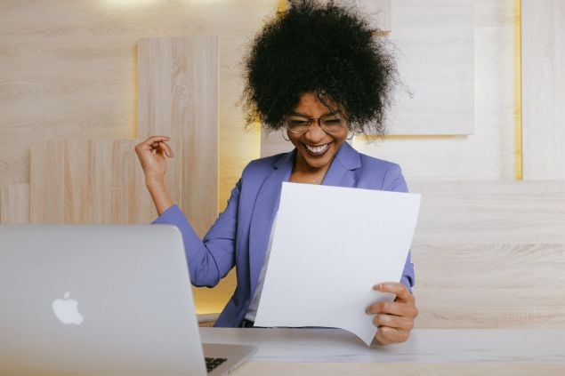 Imagem de uma mulher negra sorrindo, com roupas sociais e com papéis na mão na frente de seu laptop