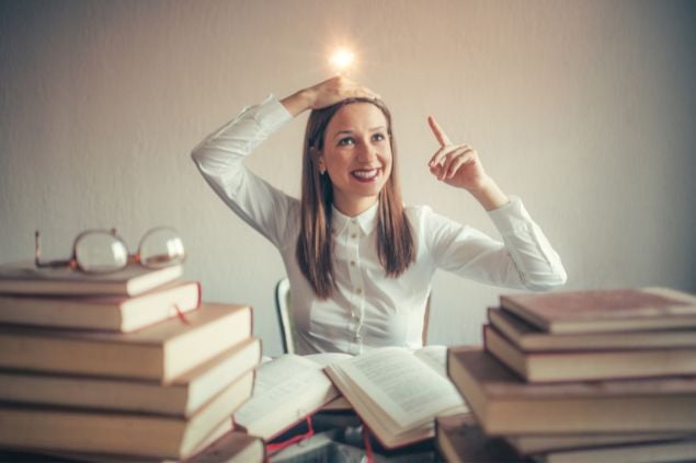 Imagem de uma mulher na frente de seus livros, com uma luz ilustrativa acima de sua cabeça. Ela está sorrindo com o dedo indicador para cima como se estivesse tendo uma brilhante ideia